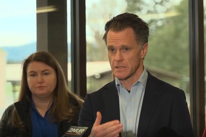 A middle-aged, grey-haired man in a dark suit gestures as he speaks to the media.