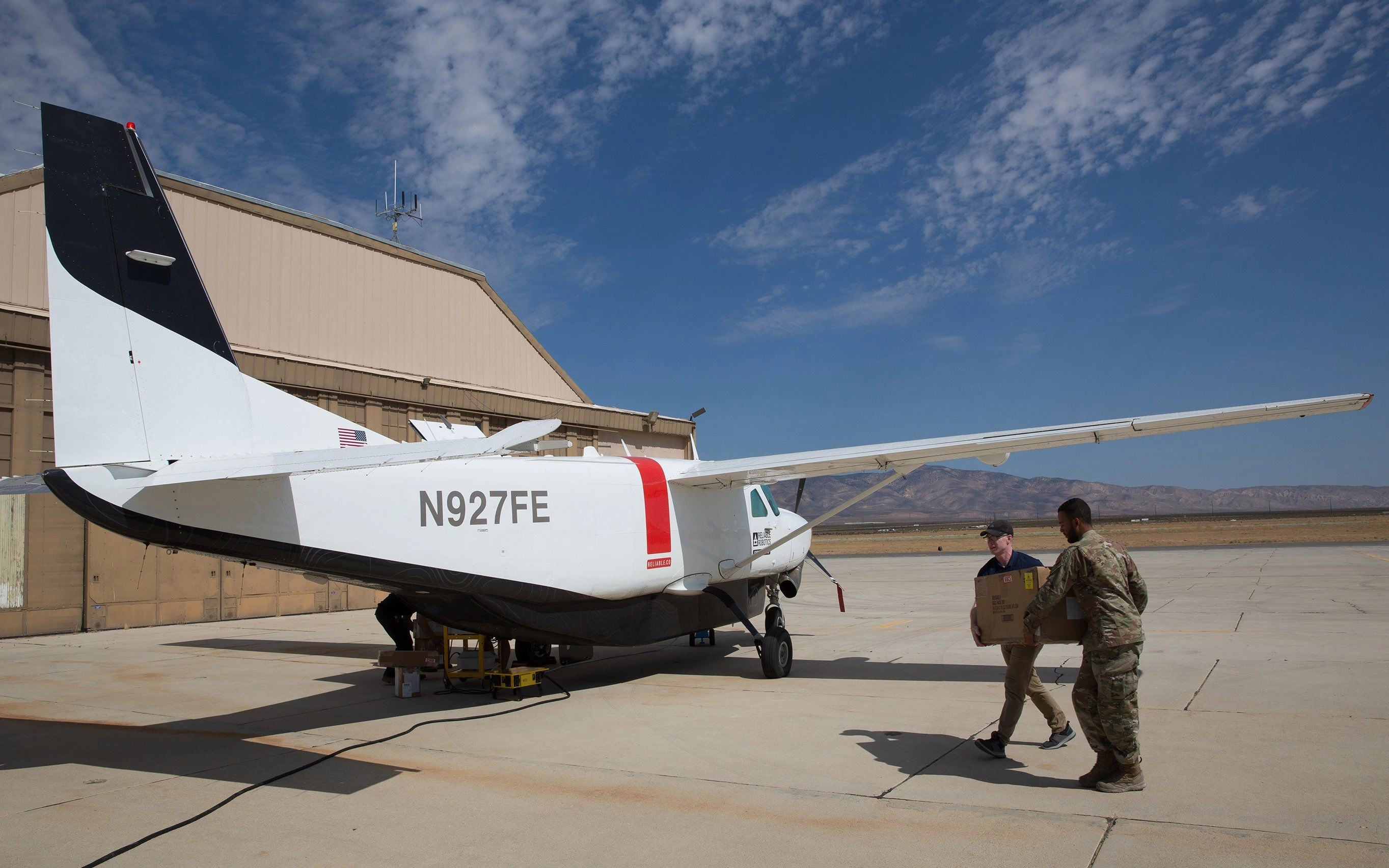 A white airplane is parked on an airport tarmac close to a hangar. Two men are carrying a large box towards the airplane.