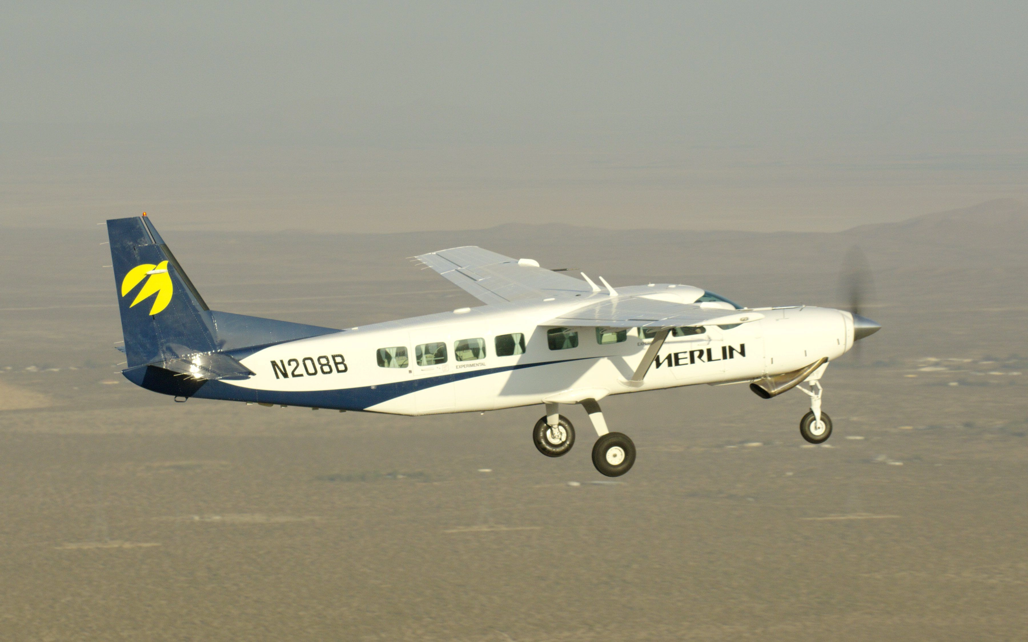 A white single-engine airplane with a high wing is seen flying over scrubby brown terrain.