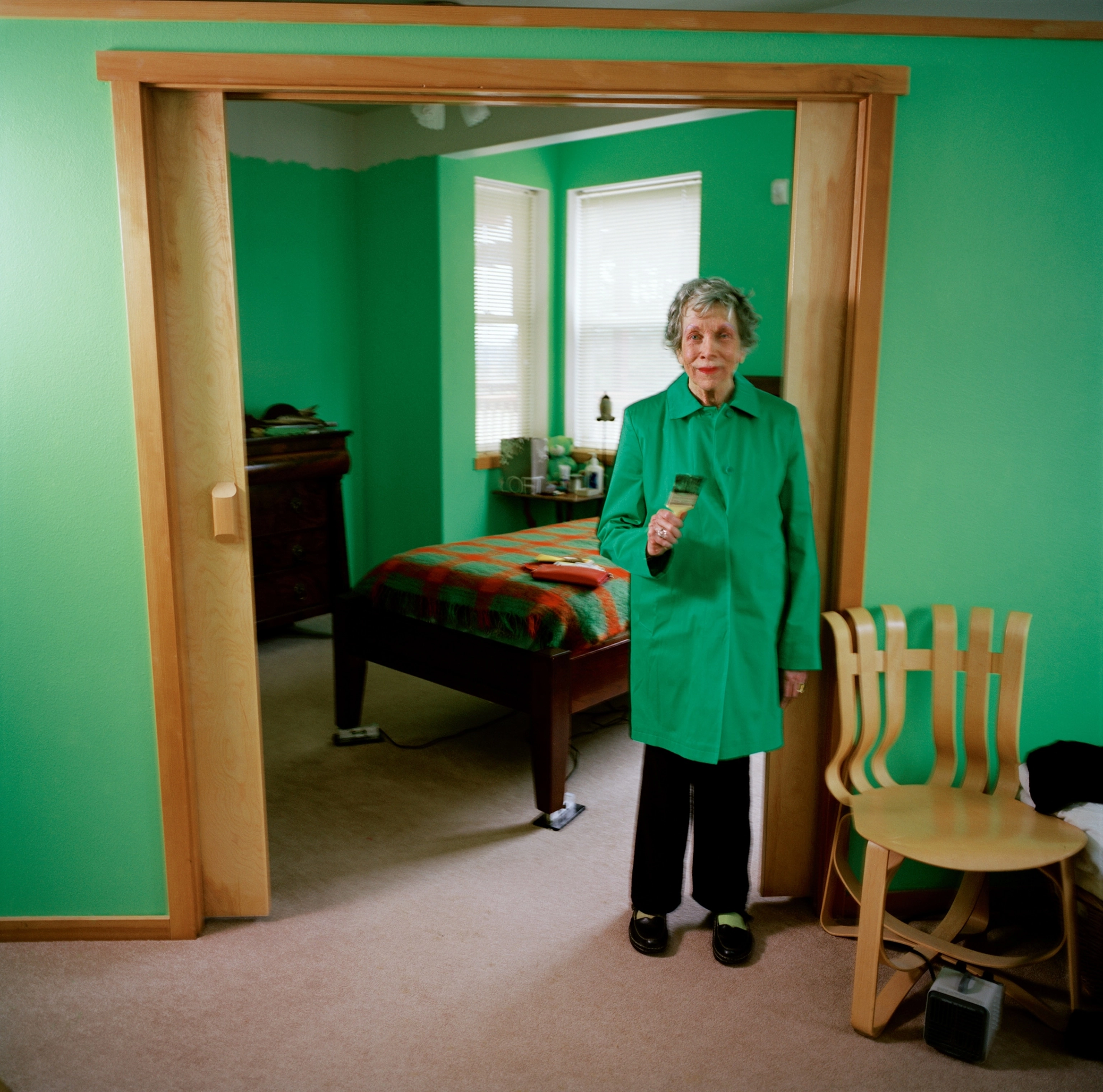An older woman stands in a green coat in a doorway. The walls surrounding her are green.