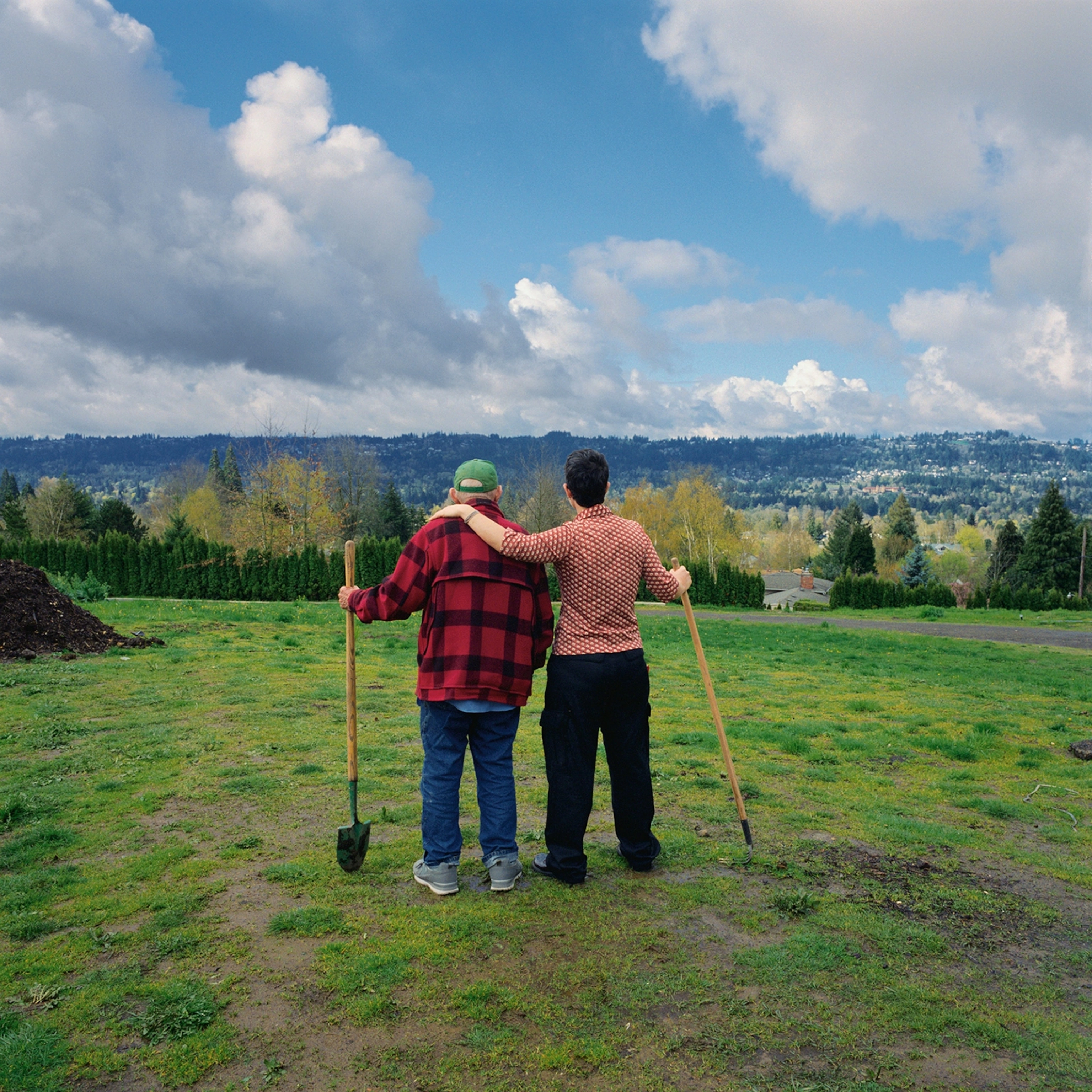 An older man and a younger man stand side by side on green grass. They are facing away from the camera and overlooking trees. The sky is blue.