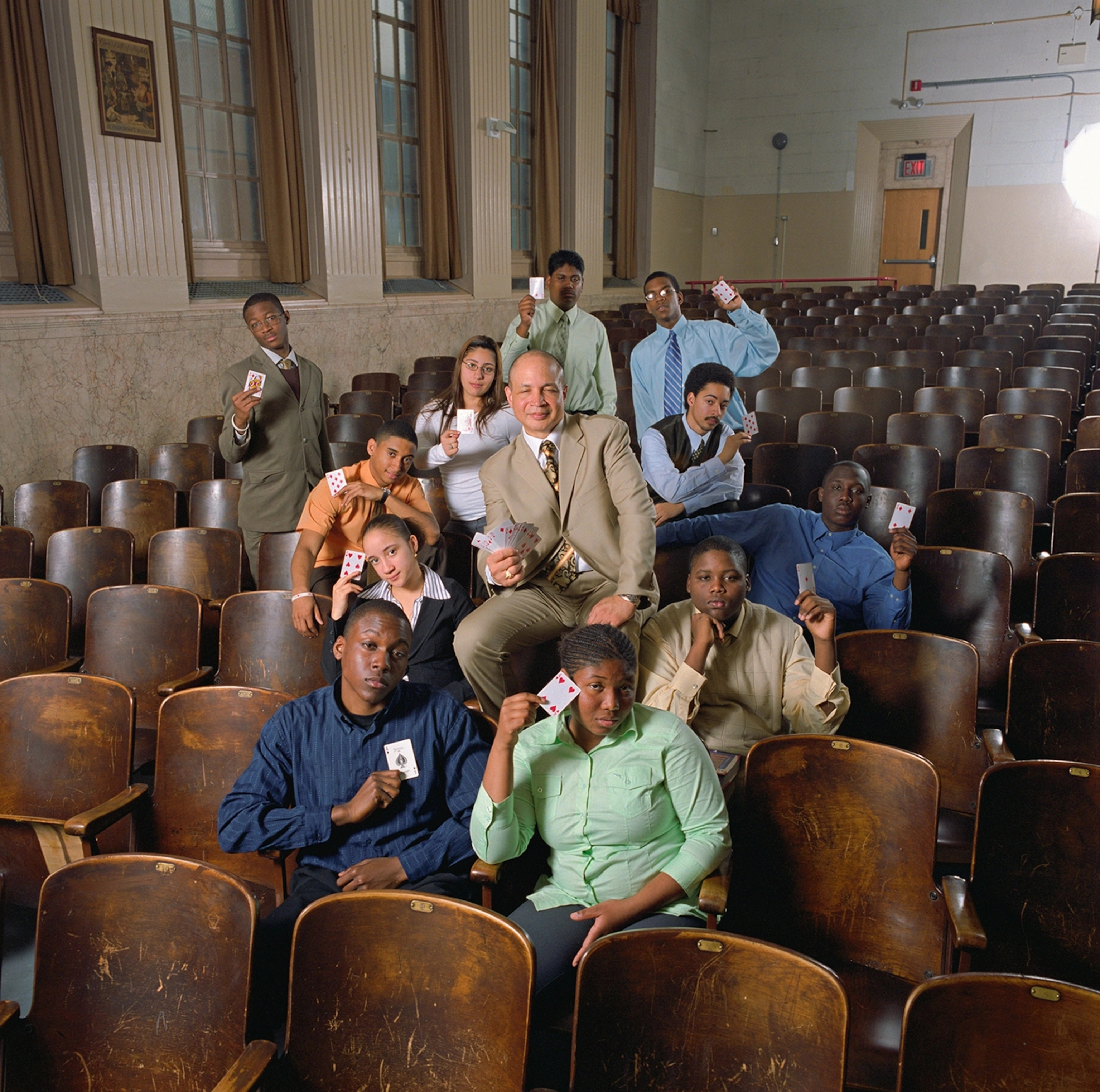 A group of people are seen in an empty auditorium, some are sitting on the brown chairs and some are standing amongst them. They are all holding playing cards.