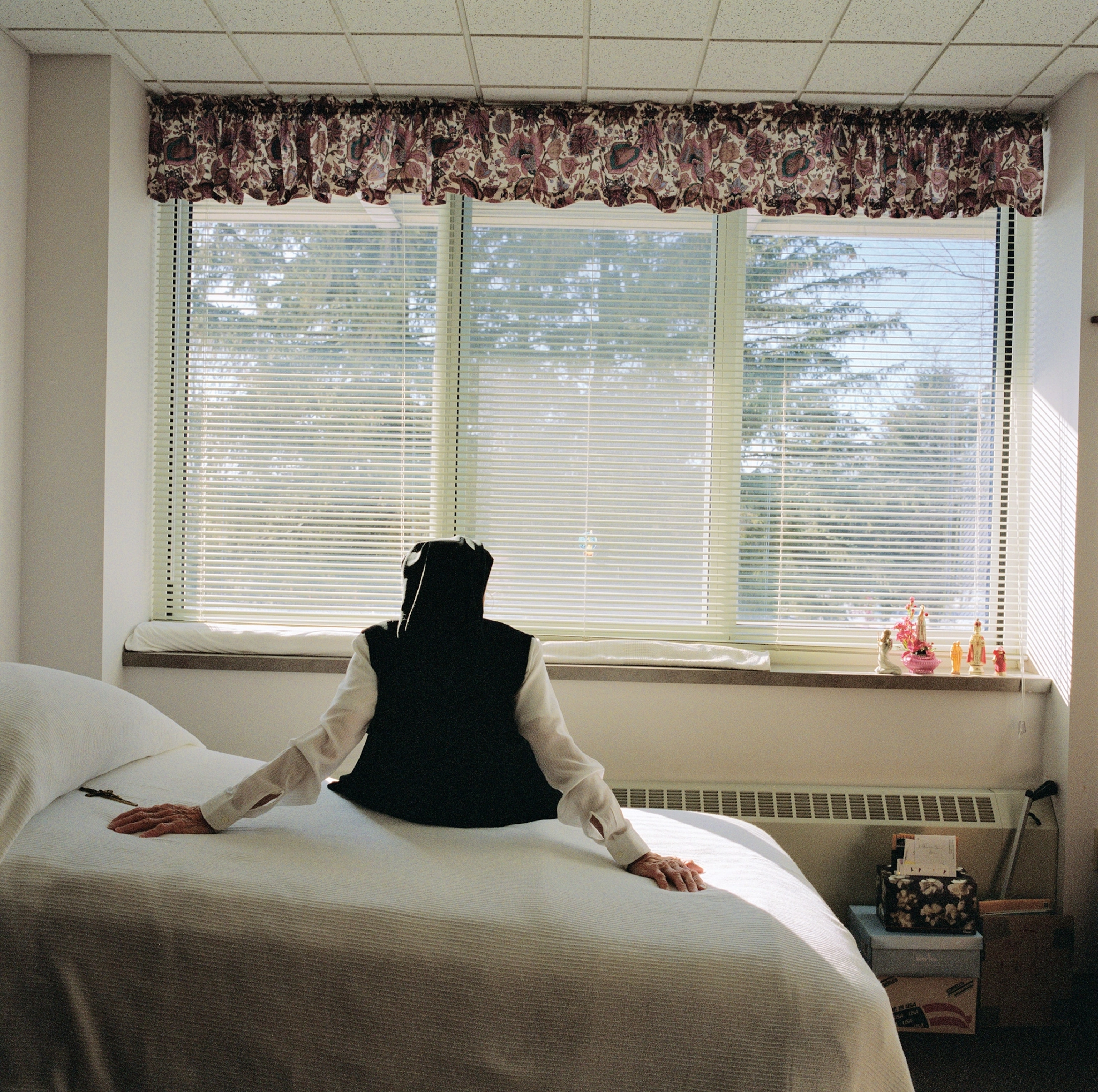 A nun sits on a twin-size bed and faces towards the window. The blinds are open so that the sunlight is seen through them.