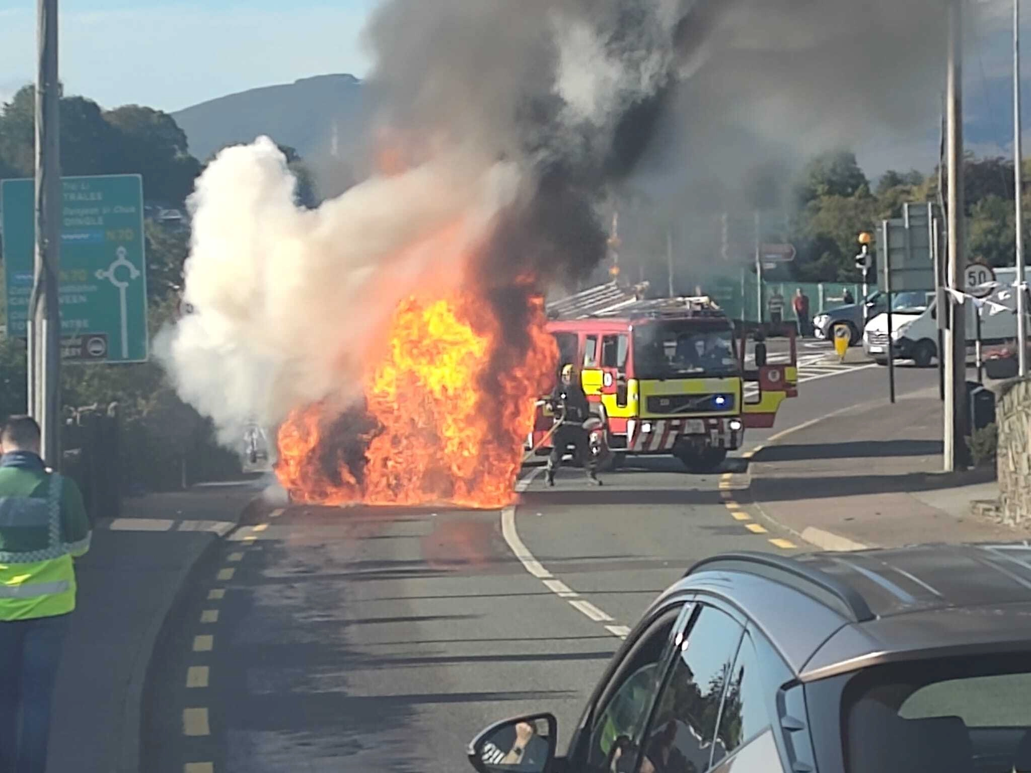 A Peugeot rental car engulfed in flames on a road, with firefighters responding.