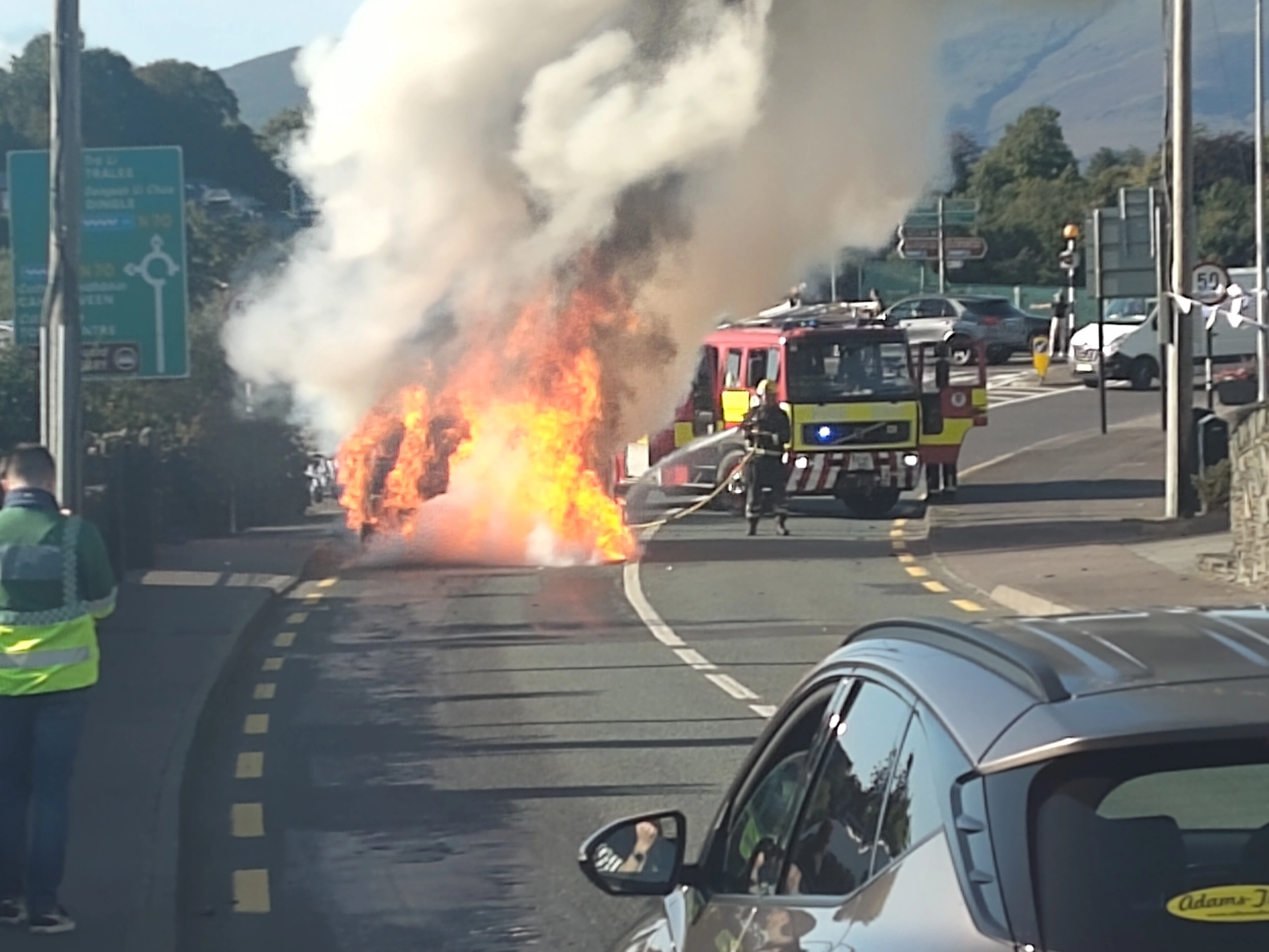 Peugeot rental car engulfed in flames on a road with firefighters responding.