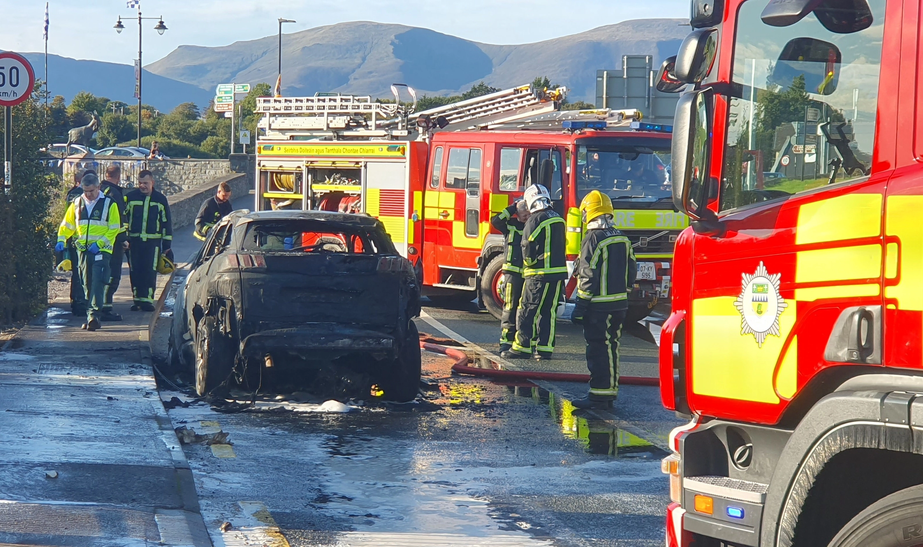 Burnt-out car on the road with firefighters.