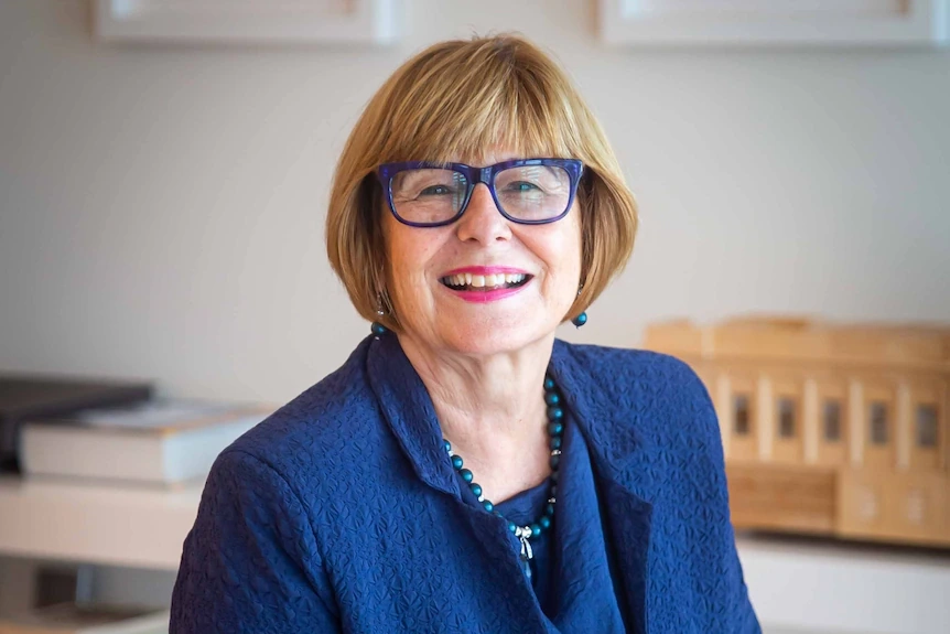 A woman with chin-length auburn hair with blue glasses and a blue suit smiles