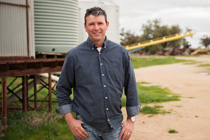 Man with white skin and blue shirt stands on rural property in front of water tank.