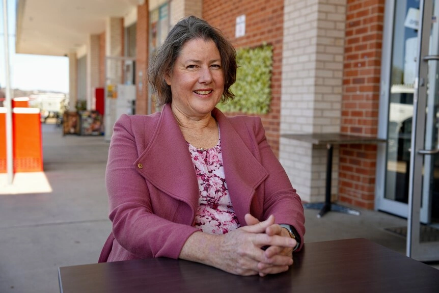 A woman with short greying brown hair sits at an outdoor table smiling.