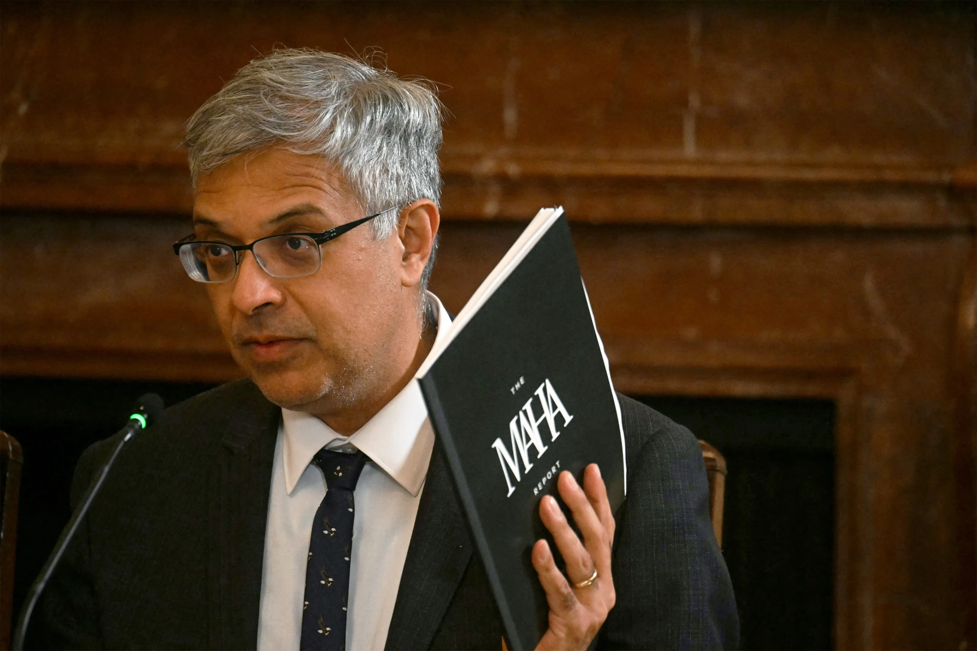 A photo of Jay Bhattacharya seated at a table, holding a printed booklet with a black cover. The cover reads, 