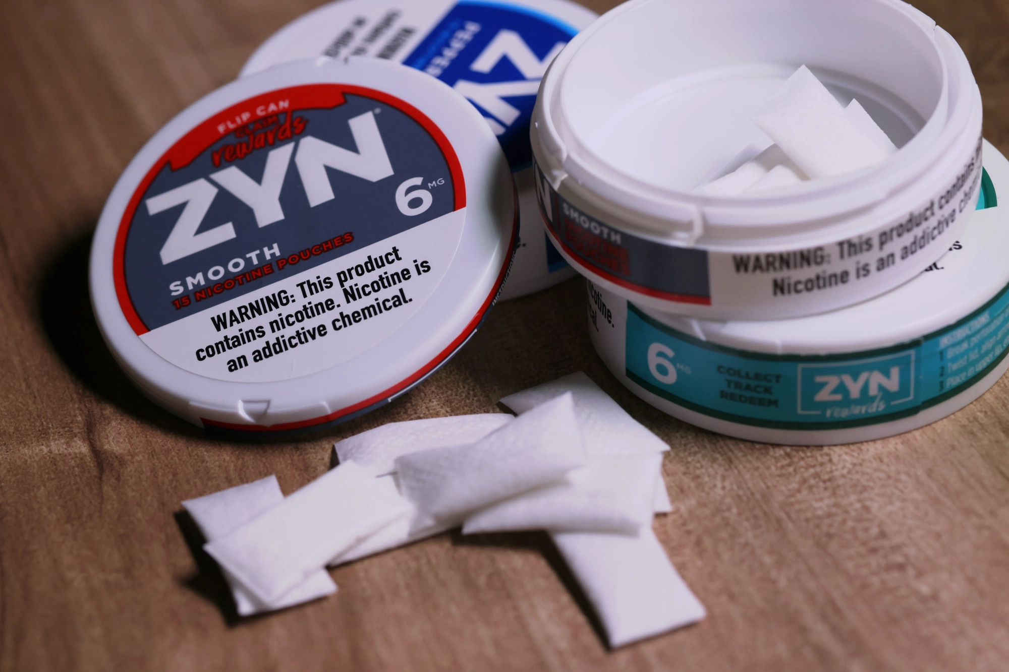 White ZYN nicotine pouch cans in red, blue, and teal labels on a wood surface, with several small white pouches spilled out, warning about nicotine's addictive chemical.