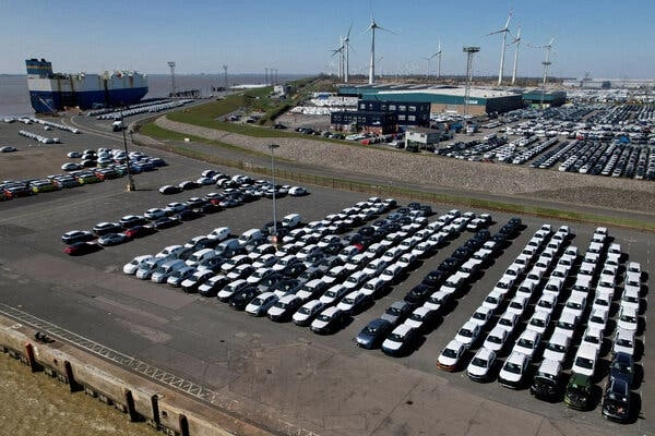 Rows of cars parked in a lot, not far from where a container ship is docked.
