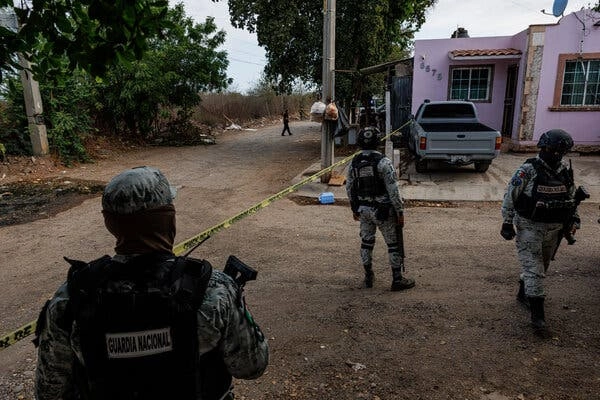 Members of the Mexican armed forces, dressed in fatigues, stand outside a home on the outskirts of Culiacan, Mexico. Yellow crime scene tape stretches across the front yard.
