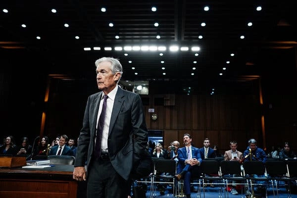 Jerome Powell stands with his right hand on a corner of a desk in a hearing room, facing away from a seated audience.