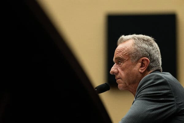 Robert F. Kennedy Jr., seen from the side in a gray suit, looks forward in front of a microphone.