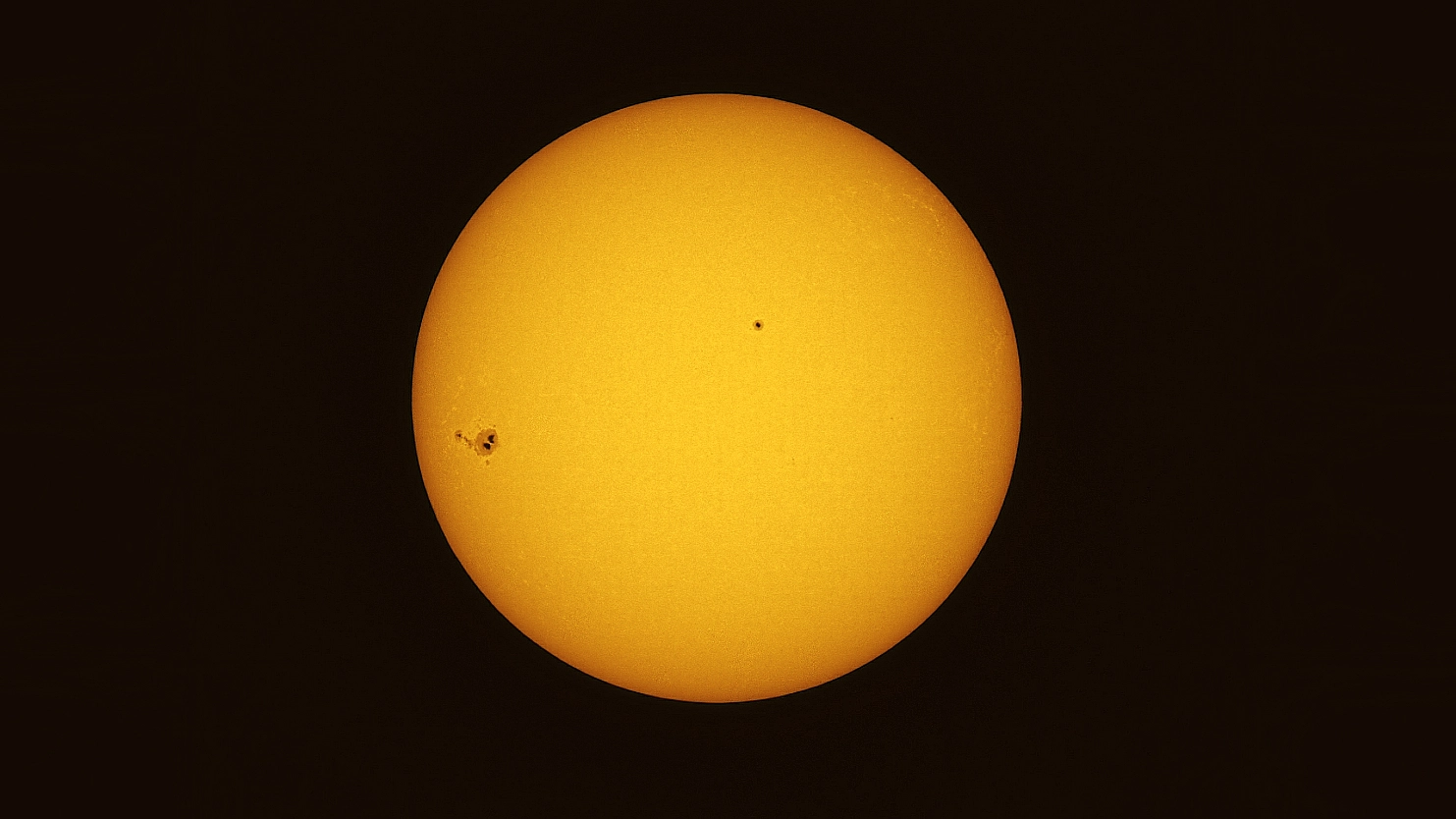 The sun's yellow disk is shown against a black background. A collection of sunspots can be seen to the left of the otherwise placid face of our star, with another close to its center.