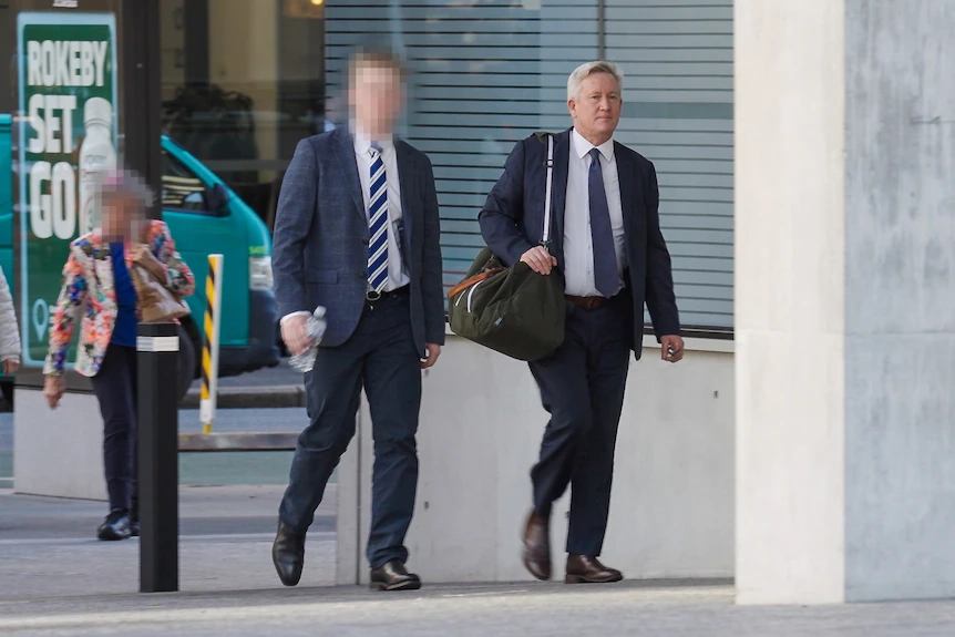 John Norman in a suit, with a bag on his shoulder walking in the Brisbane District Court, 25 June 2025.