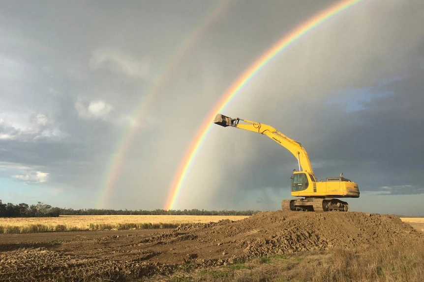 A yellow excavator with the bucket raised, on a mound of dirt, against a rainbow, near Toobeah, QLD, date unknown.