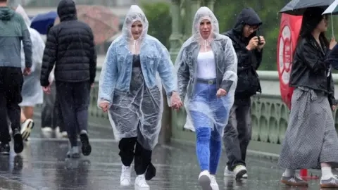 PA Media Two women in plastic overalls walk in the rain across a bridge in London.
