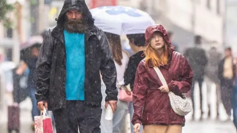 PA Media A man and woman walk along a street in Leeds, wearing rain jackets with hoods up as rain pours down.