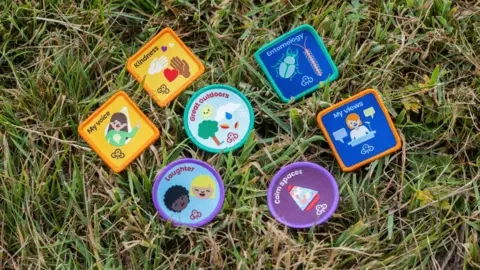 Girlguiding Eight of the new badges laid out on the grass. They are: my voice, kindness, laughter, great outdoors, entomology, clam spaces and my views.