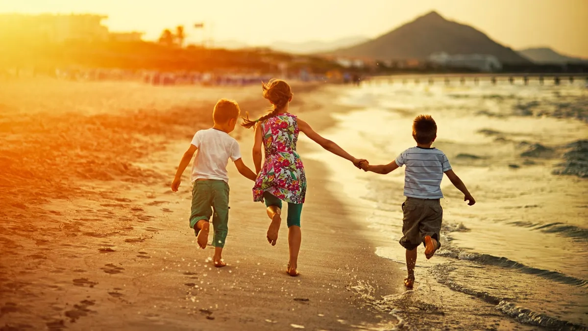 Three kids seen from behind running down beach at sunset holding hands