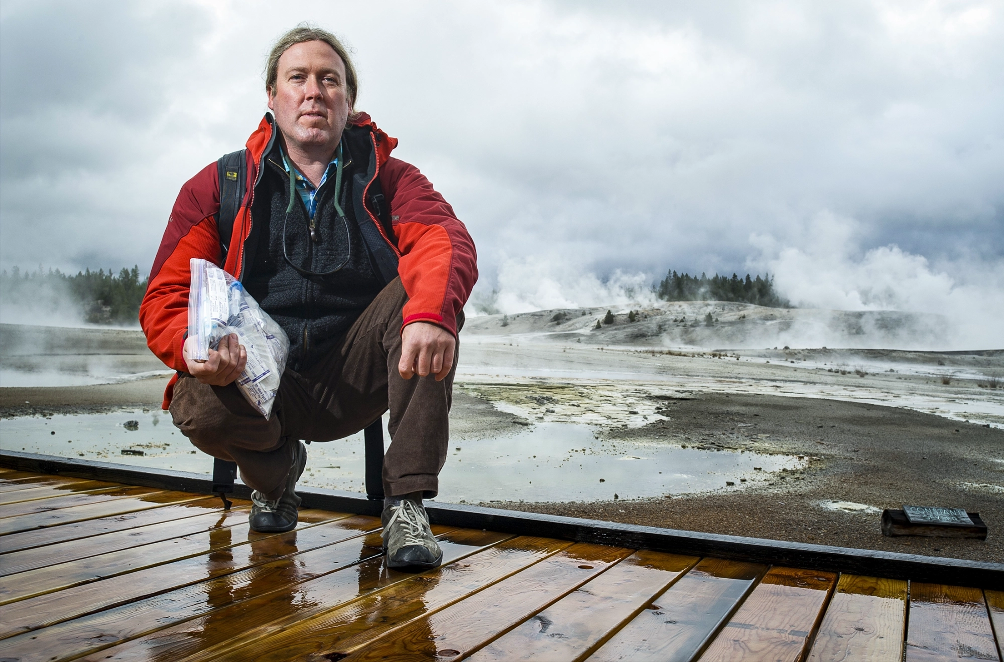 Eric Boyd stands on a boardwalk holding a bag of microbial samples collected from the hot spring behind him.