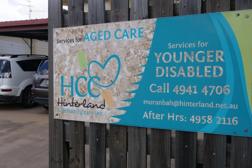 A sign reading 'Hinterland Community Care', affixed to a wood fence, with two SUV cars in the background.