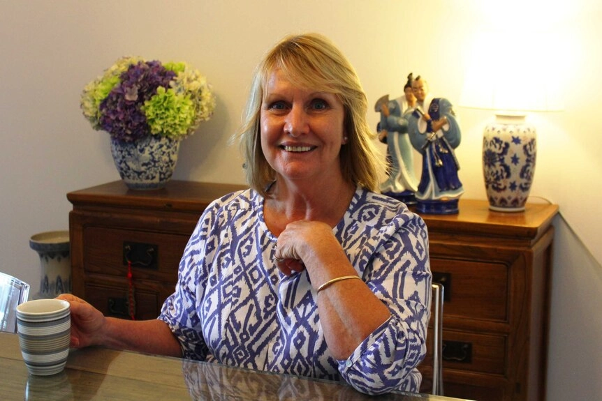 A blond woman, sitting at her kitchen table with a cup of tea.