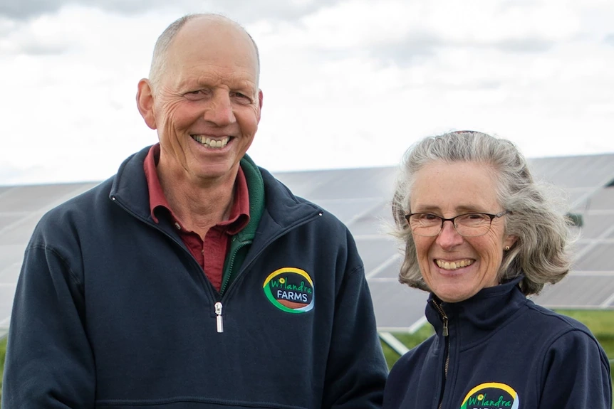 A middle-aged man and woman stand outside in front of several solar panels, smiling at the camera