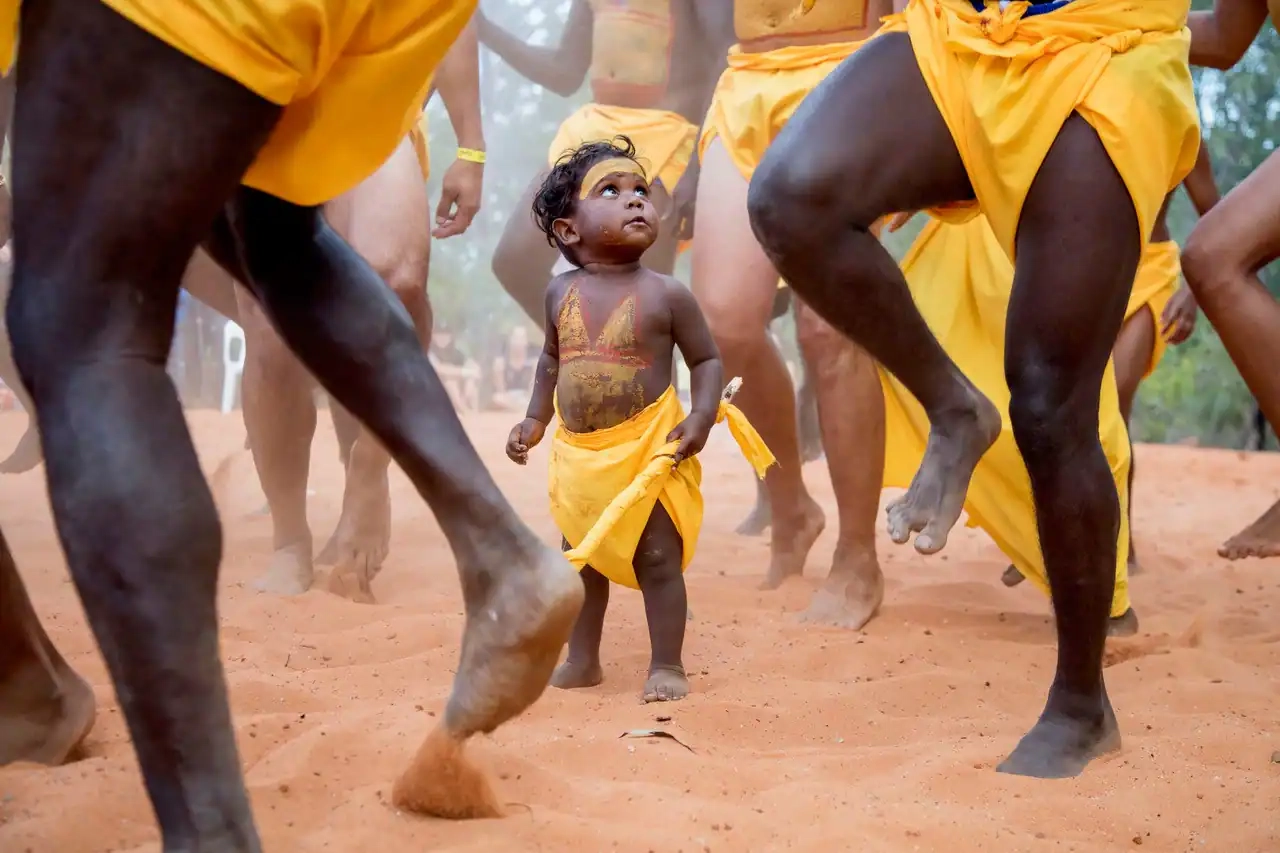 A young child in a yellow garb tied around his legs stands in the centre, looking at adult dancers during a traditional dance performance. 