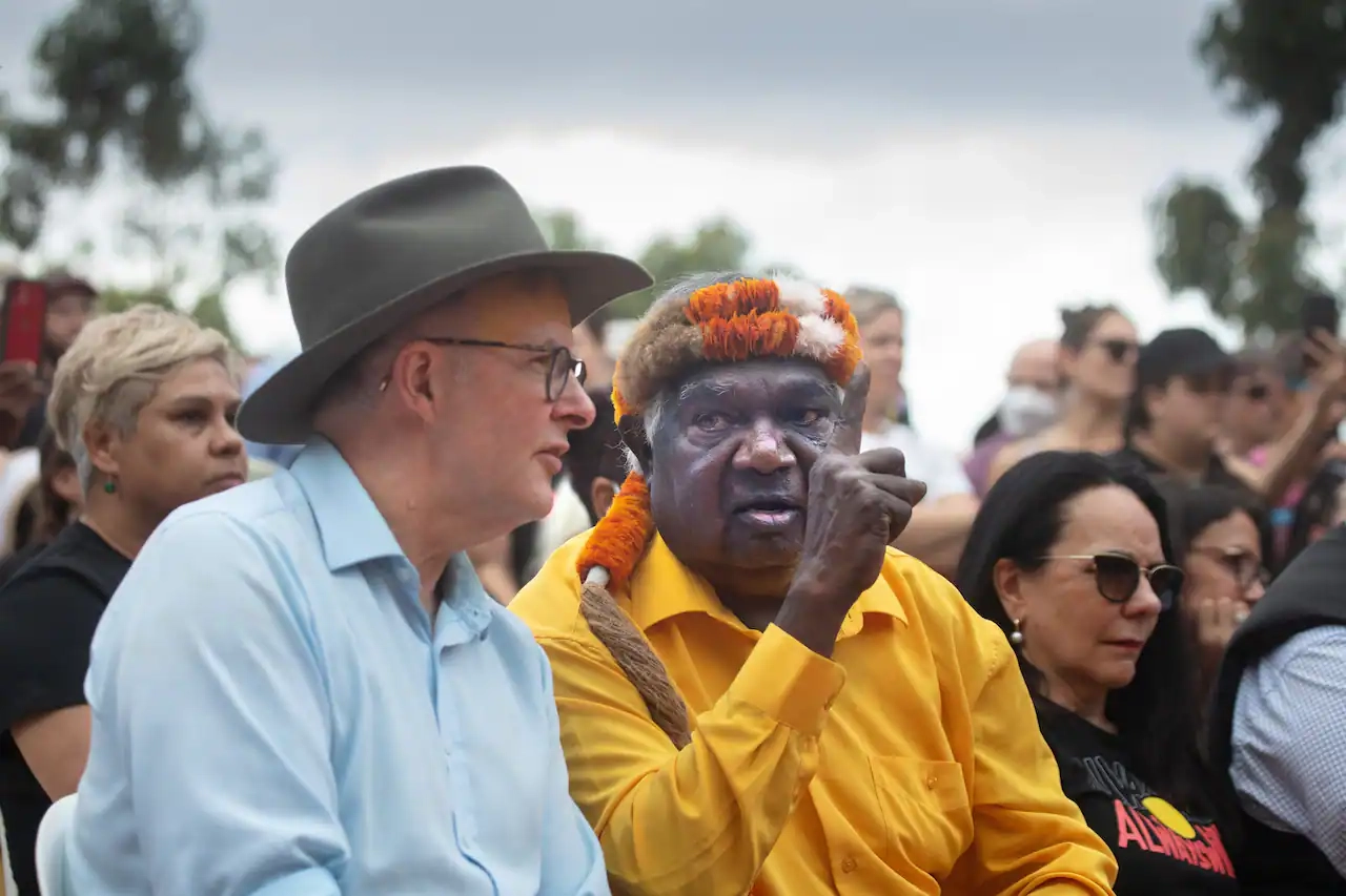 Anthony Albanese, wearing a hat, is sitting next to a man in a yellow shirt who is speaking with his finger pointing upwards. There's a crowd sitting around them.