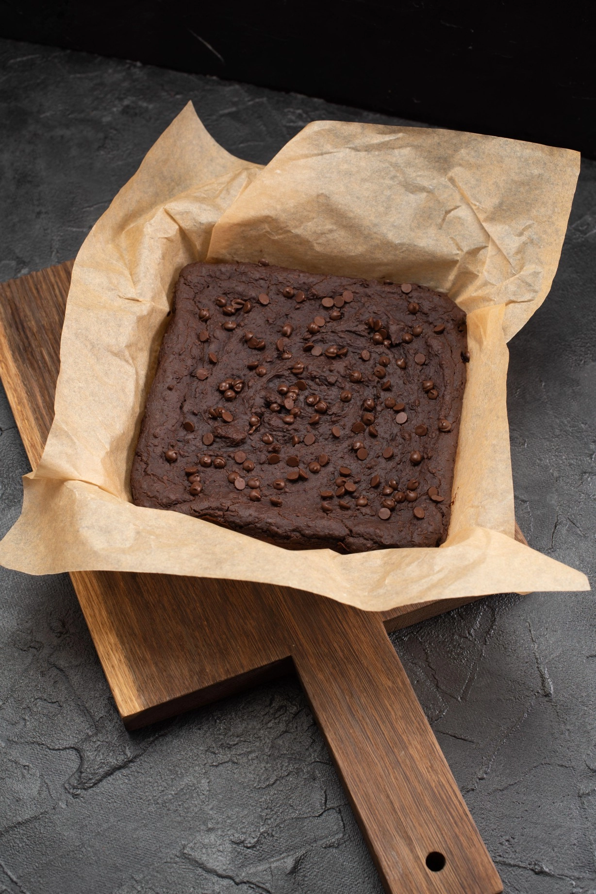 2F2AEKC Uncutted brownies with choco drops in baking paper on dark plaster background high angle view