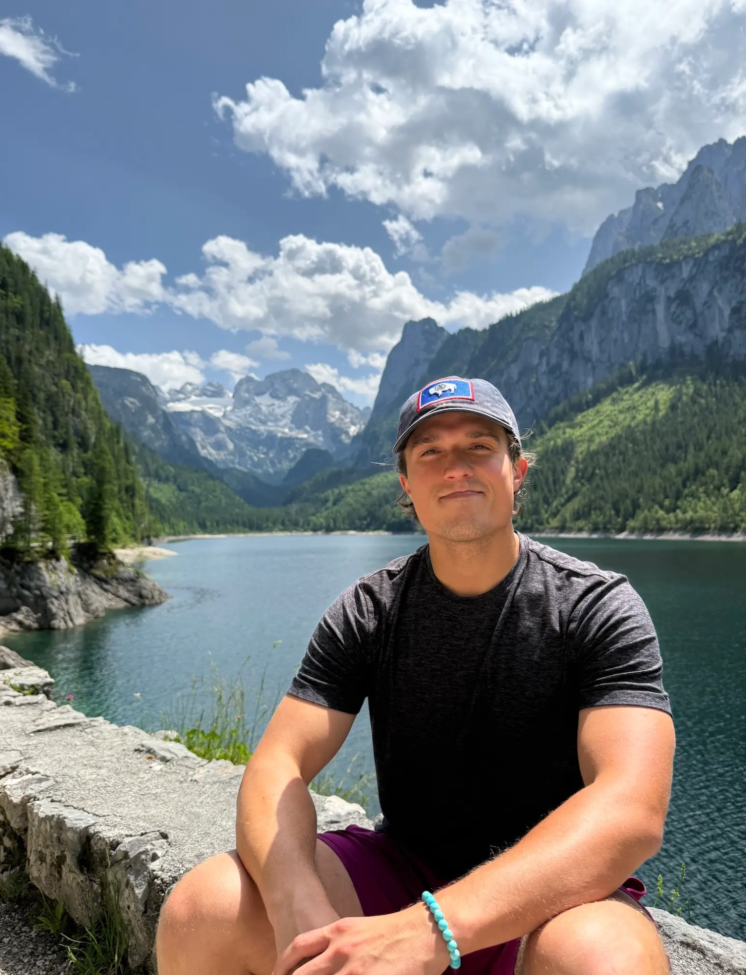 A young man in a baseball cap and t-shirt poses in front of beautiful mountains and forest