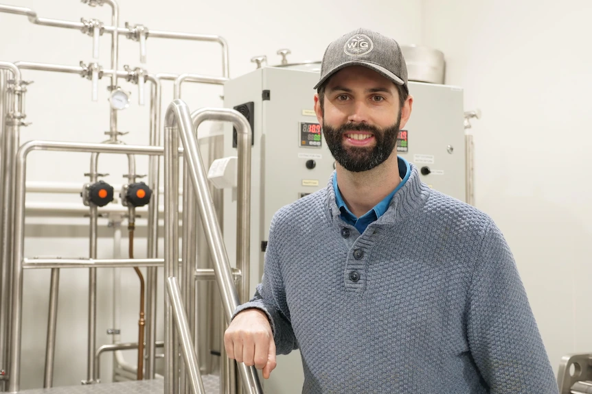 A man in a cap and grey knitted jumper smiling at the camera with brewery equipment in the background. 