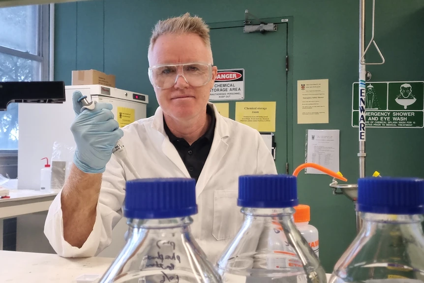 A man in a laboratory wearing a white jacket and safety glasses with a device in his hand