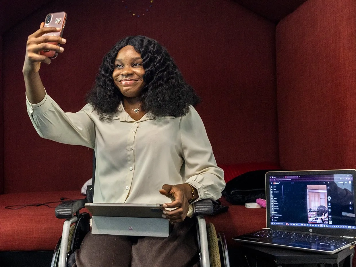 Photo of a smiling woman in a wheelchair taking a selfie, with an open laptop at her side.
