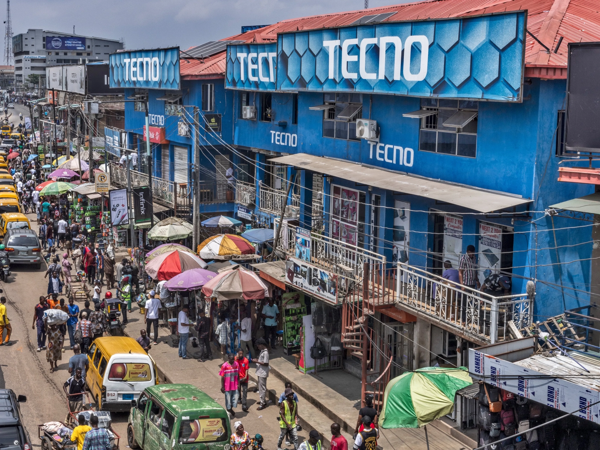 Photo of a crowded city street, with colorful cars and umbrellas, and the word Tecno on several blue buildings.