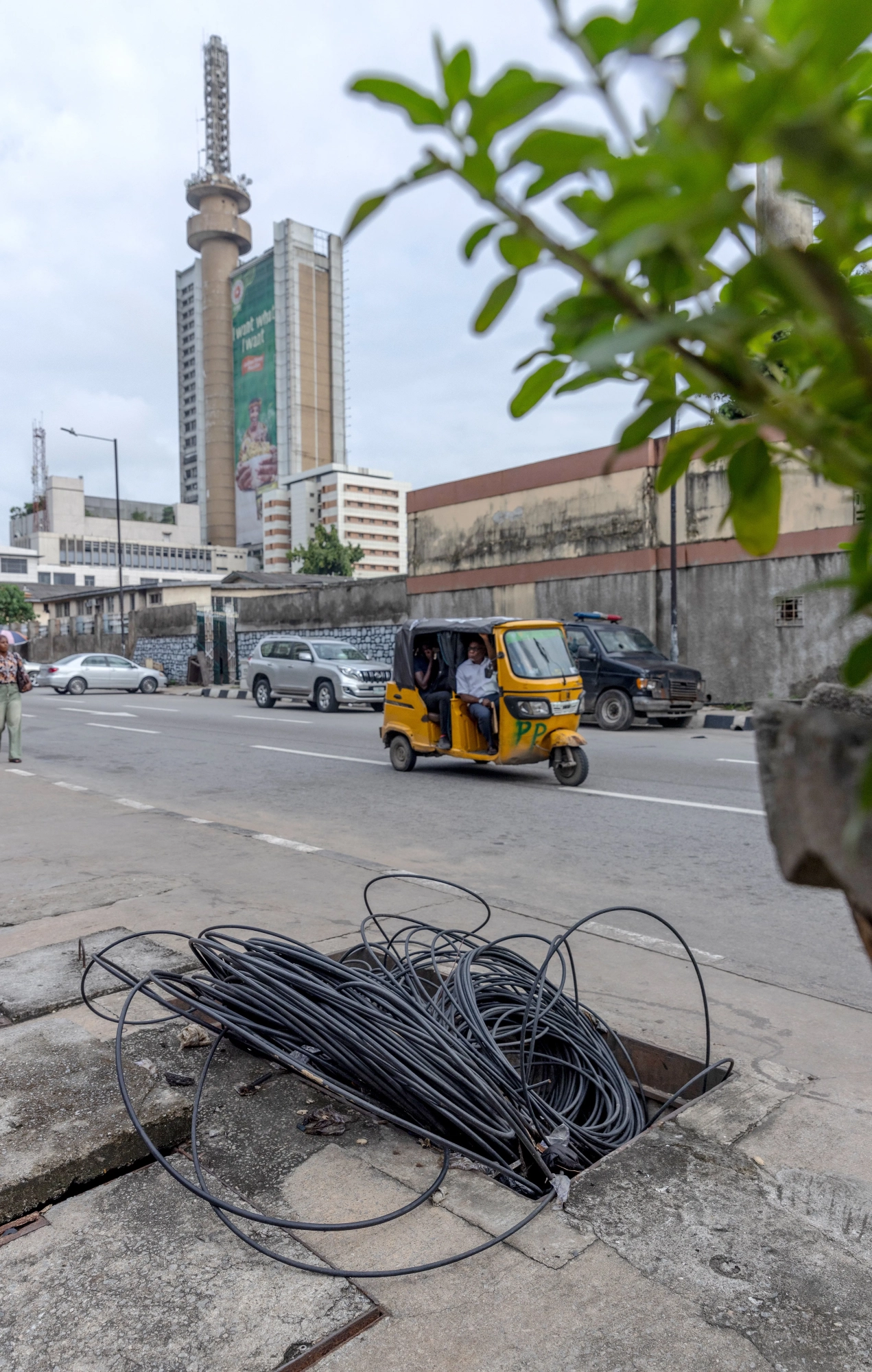 Photo of tangled black cables spilling from a rectangular opening in the sidewalk, with street traffic and a distinctive tall building in the background.
