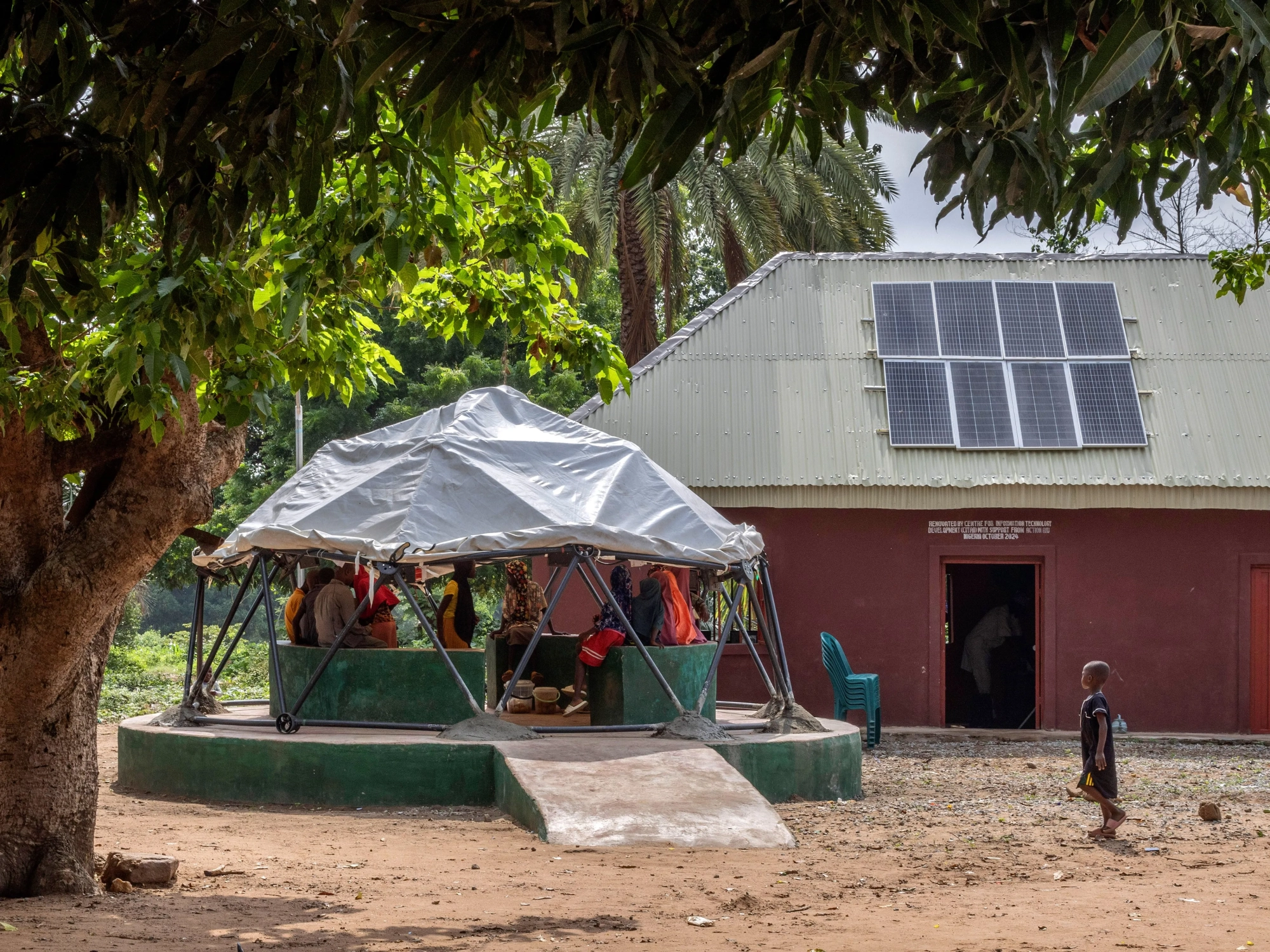 Photo of a plastic tarp covering a round green structure on a platform, with a group of people gathered inside. A building with solar panels is in the background.