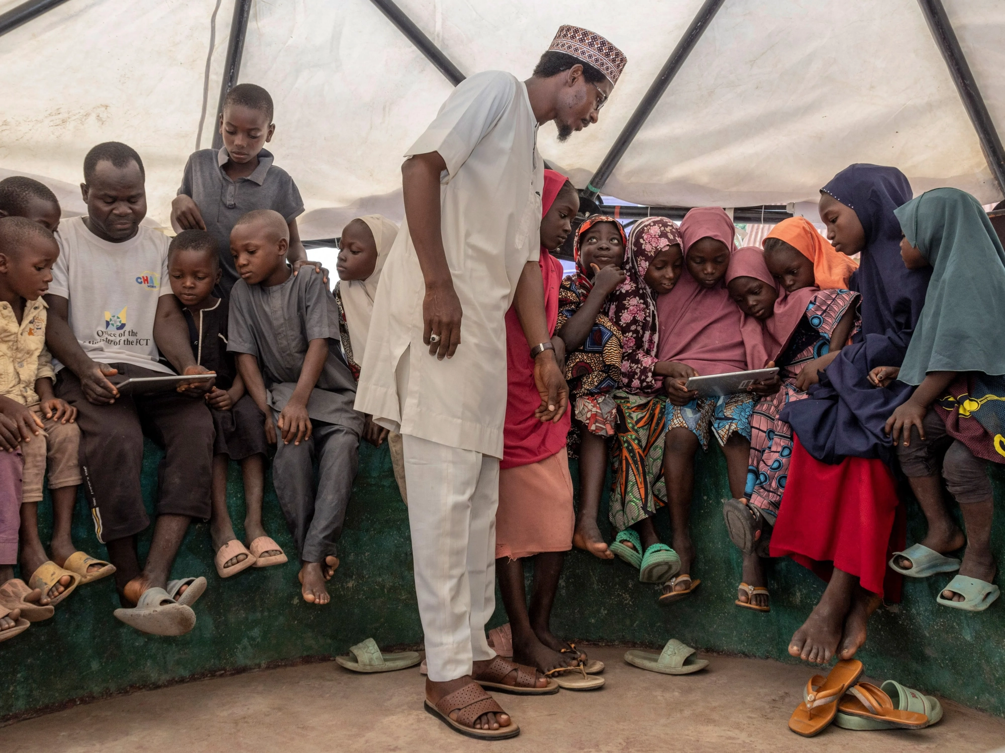 Photo of a man in Nigerian garb standing in front of a seated group of mainly children who are looking at tablets.