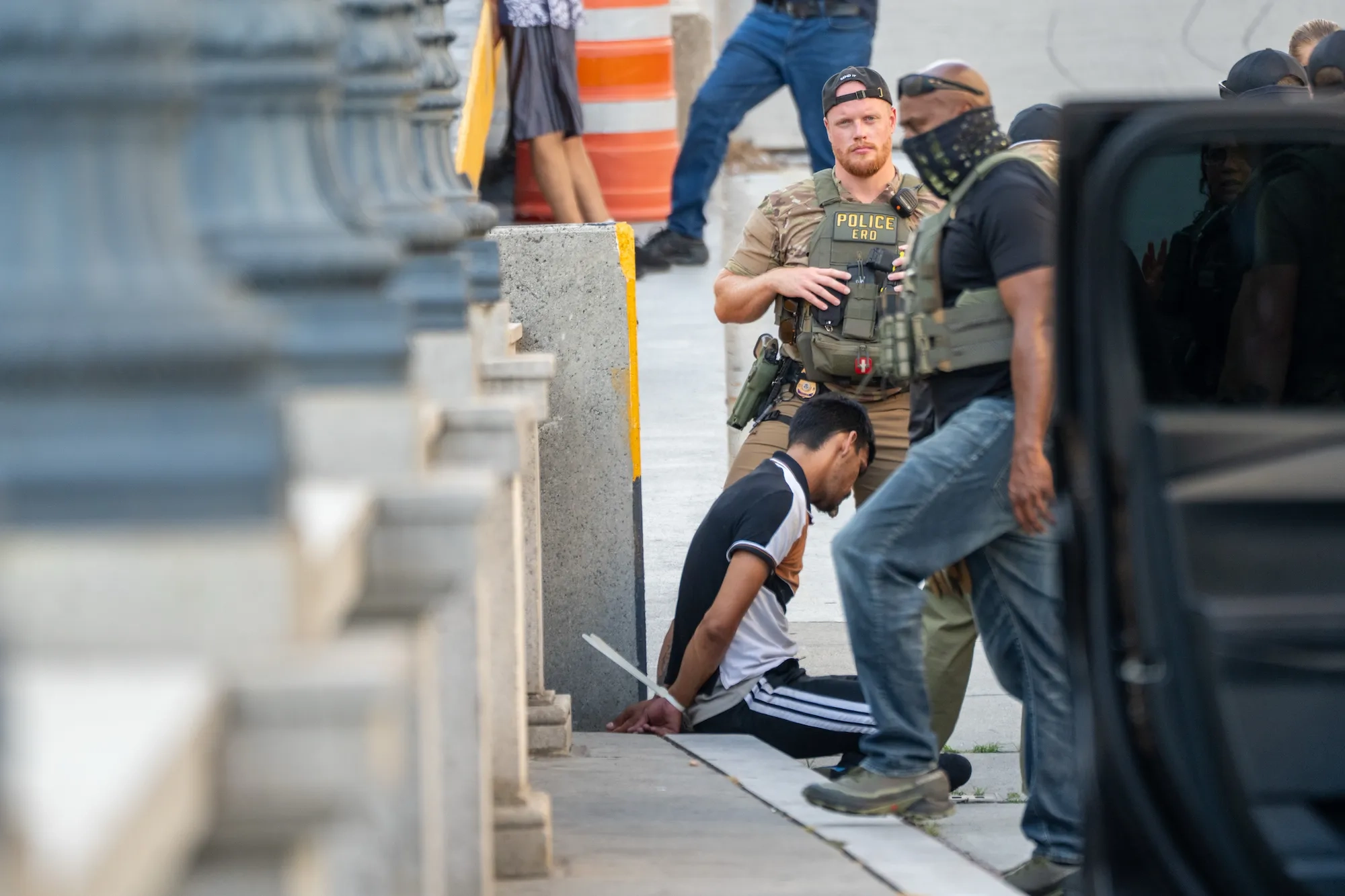 A man in a black and white shirt sits handcuffed on the sidewalk near concrete barriers while police officers, one wearing a vest labeled 