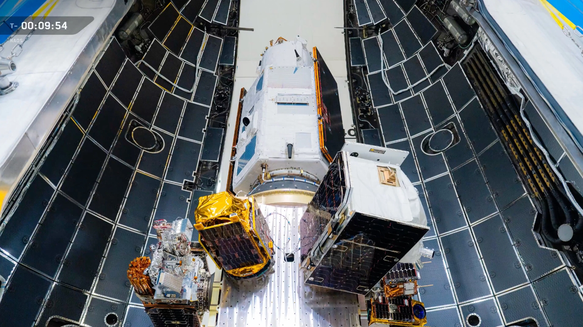 a handful of boxy metallic satellites are seen attached to a rocket's upper stage inside a hangar